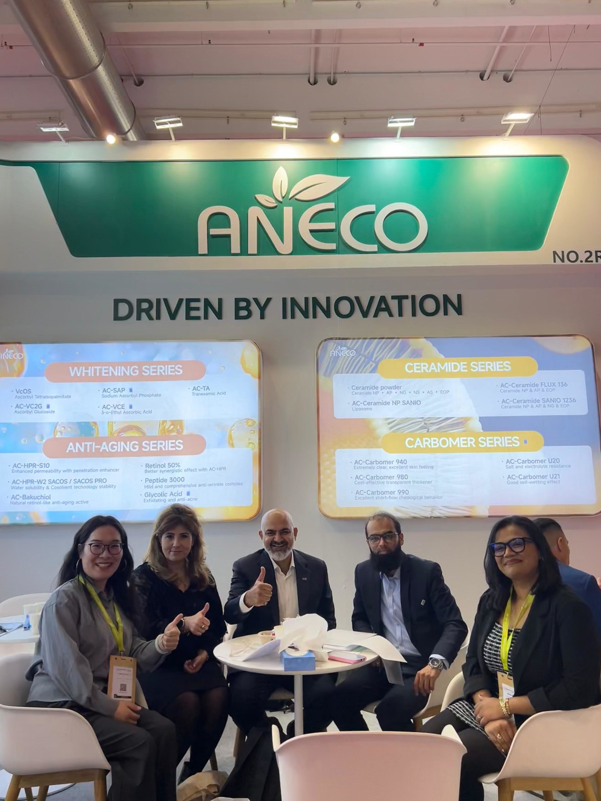 Five professionals seated around a white table at an ANECO booth, with a 'Driven by Innovation' banner and product boards in the background.