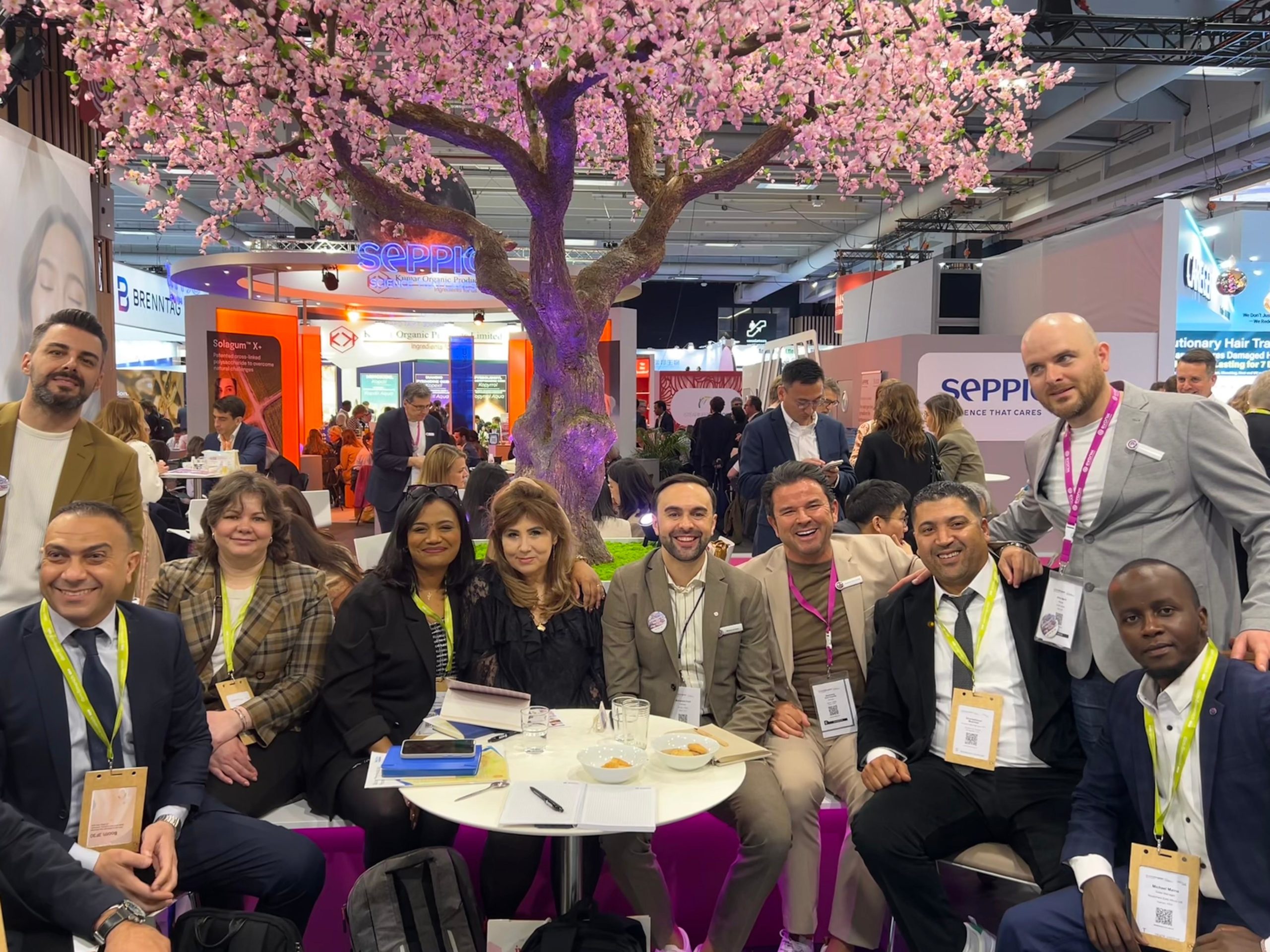 Group of professionals at a trade show seated around a round table under a pink blossom tree in the exhibition hall.
