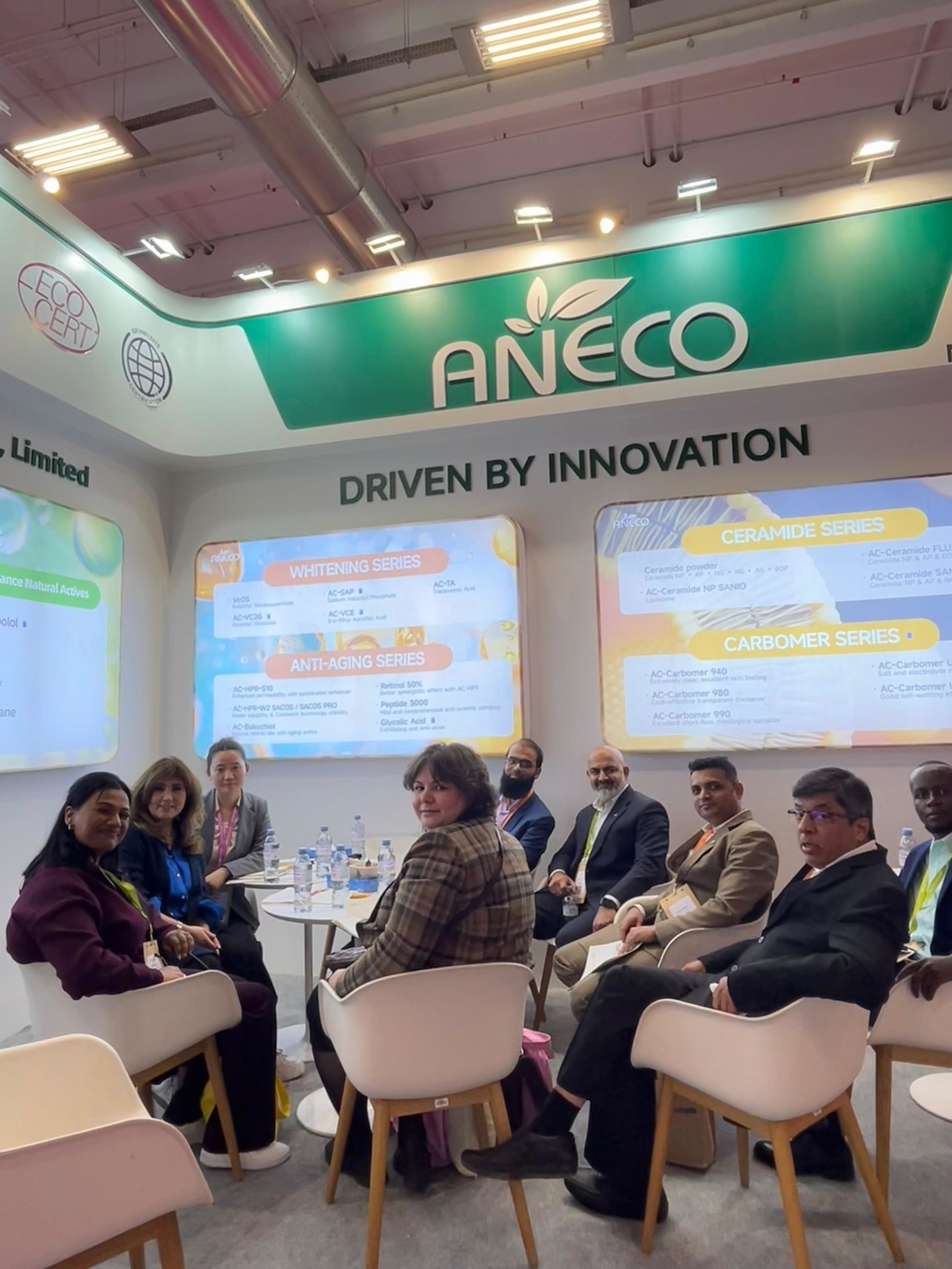 Group of professionals seated around tables at a trade show booth with ANECO branding in green on the backdrop behind them.
