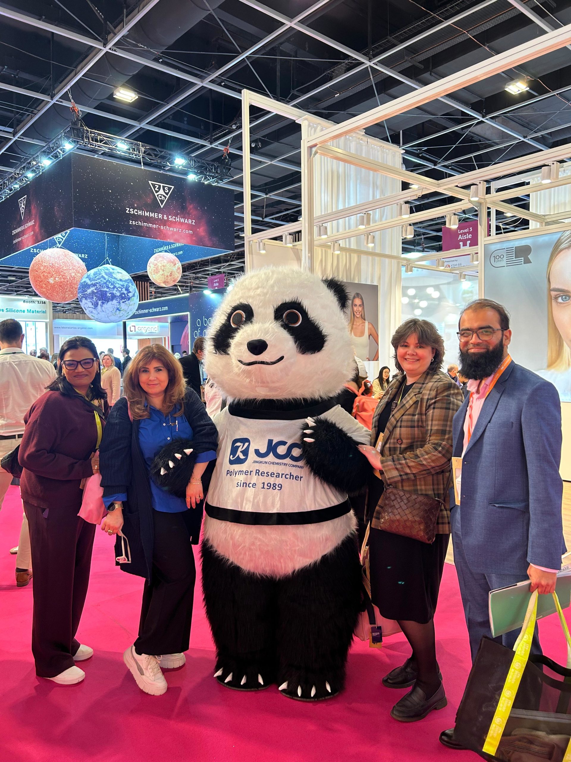 Group of people posing with a panda mascot at a trade show, pink carpet and exhibition booths in the background.