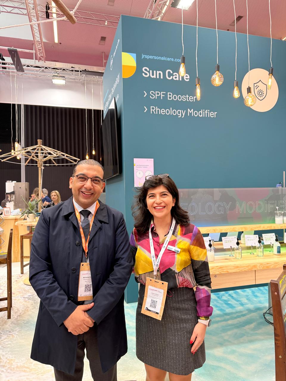 Two smiling professionals stand at a teal trade show booth with Sun Care branding and product names like SPF Booster and Rheology Modifier.