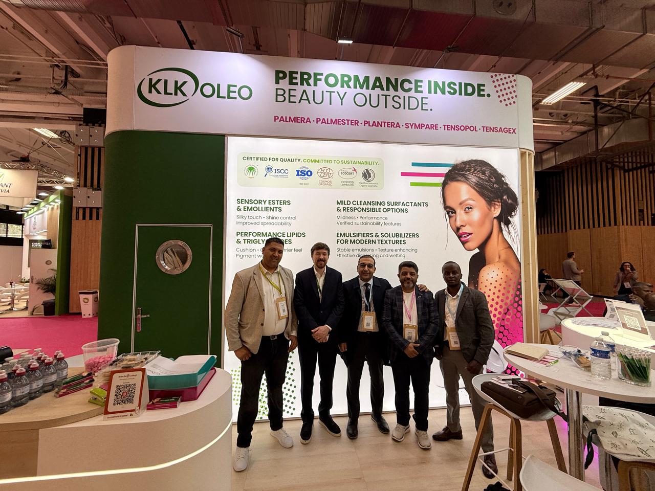 Five men in suits pose together at a trade show booth for KLK OLEO, with a large banner reading 'Performance Inside. Beauty Outside'.