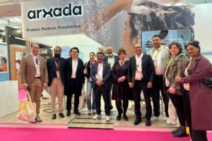 Group of professionals posing at an exhibition booth with a large arxada banner and pink carpet.