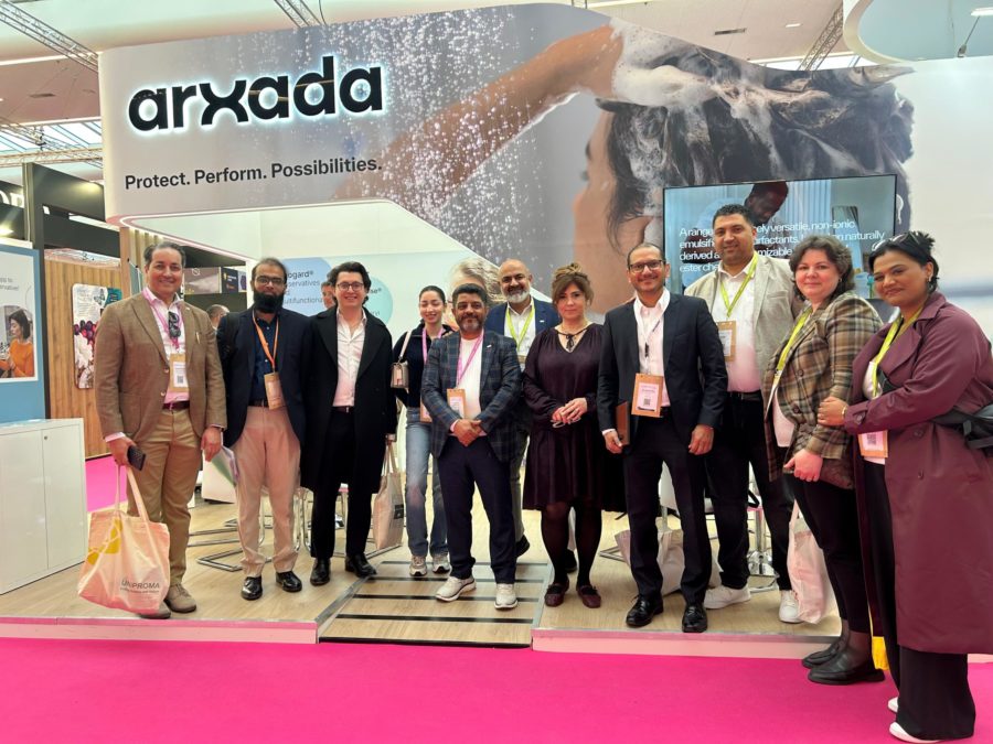 Group of professionals posing at an exhibition booth with a large arxada banner and pink carpet.