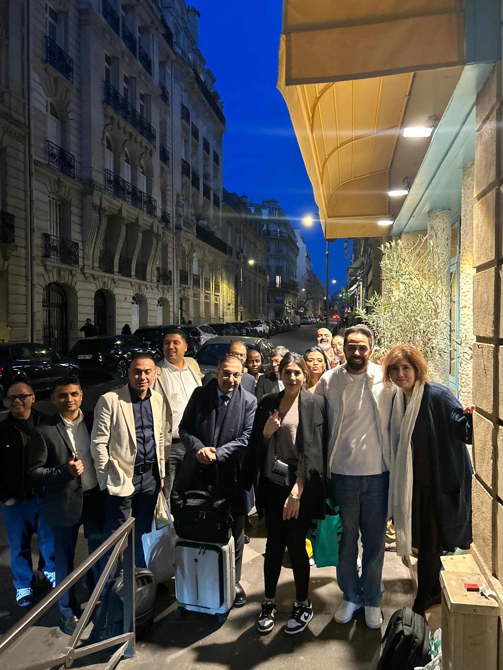 Group of people posing on a city sidewalk with luggage in front of a storefront at dusk.