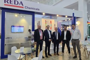Five men in suits pose at the REDA Chemicals booth during a trade show, with blue framework and glass tables nearby.