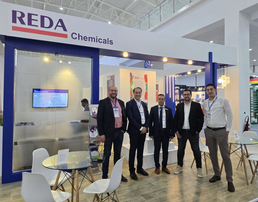 Five men in suits pose at the REDA Chemicals booth during a trade show, with blue framework and glass tables nearby.
