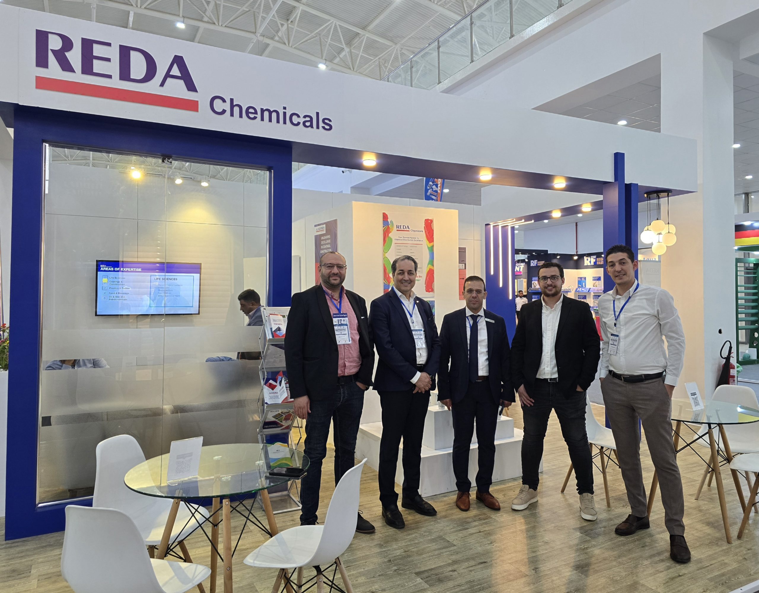 Group of five men in business attire standing at a REDA Chemicals booth at a trade show, posing for the photo.
