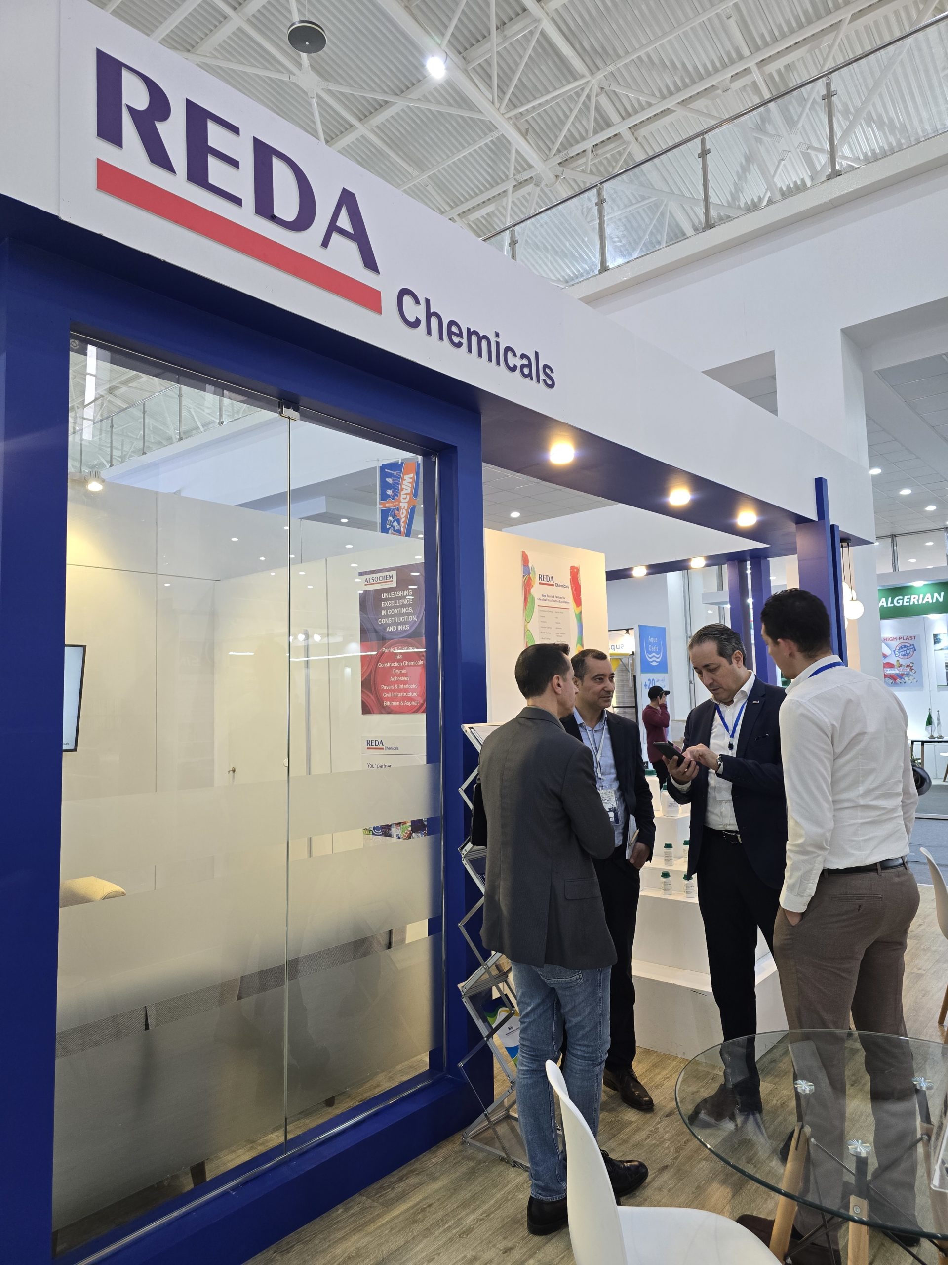 Trade show booth with a large 'REDA Chemicals' sign in purple and red; three men in suits talk near the glass wall.
