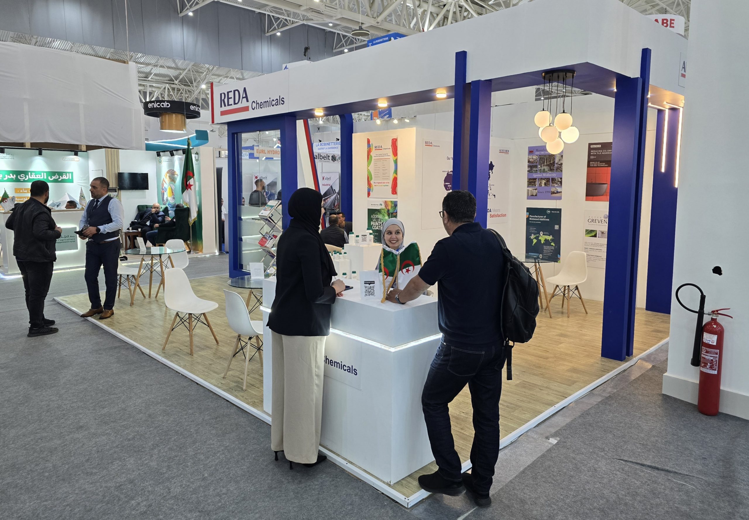 Trade show booth for REDA Chemicals with blue framework, people talking at a white counter and posters in the background.