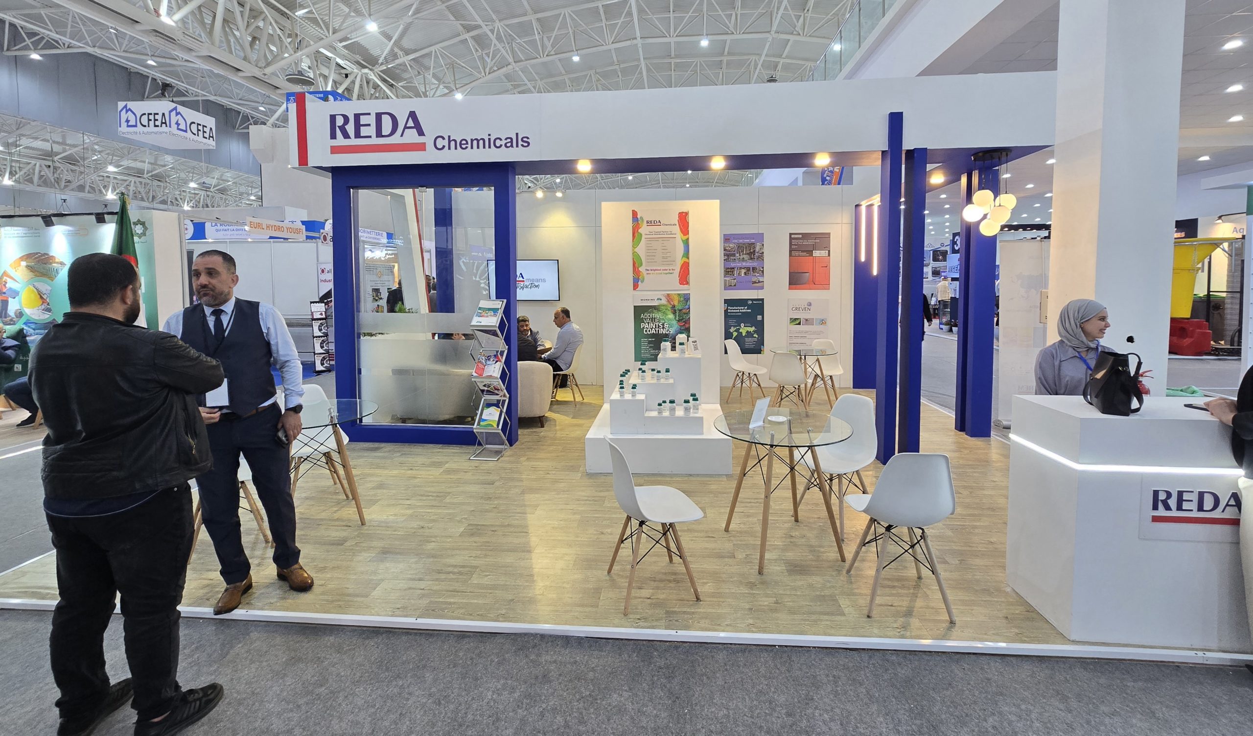 Booth for REDA Chemicals at a trade show with white and blue pillars, seating area, and posters on display walls.