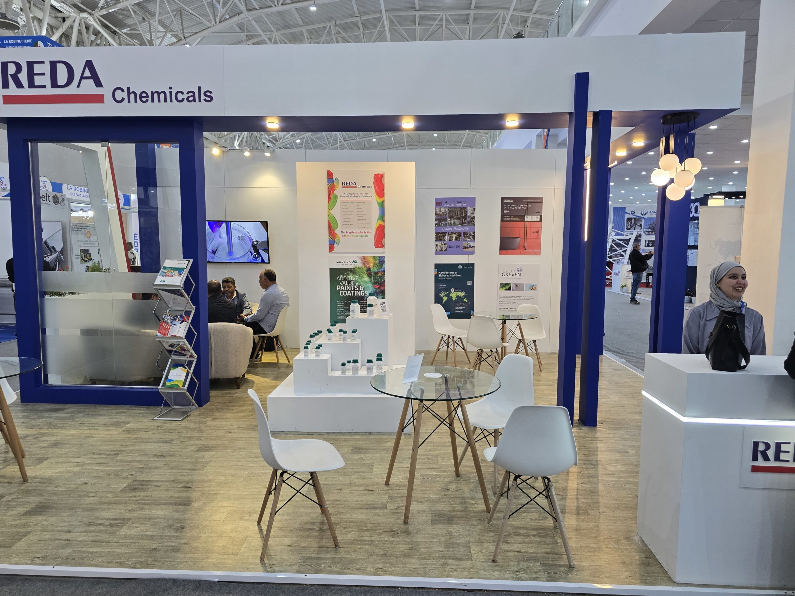 Trade show booth for REDA Chemicals with white walls, blue framing, posters, and white seating around glass tables.",
