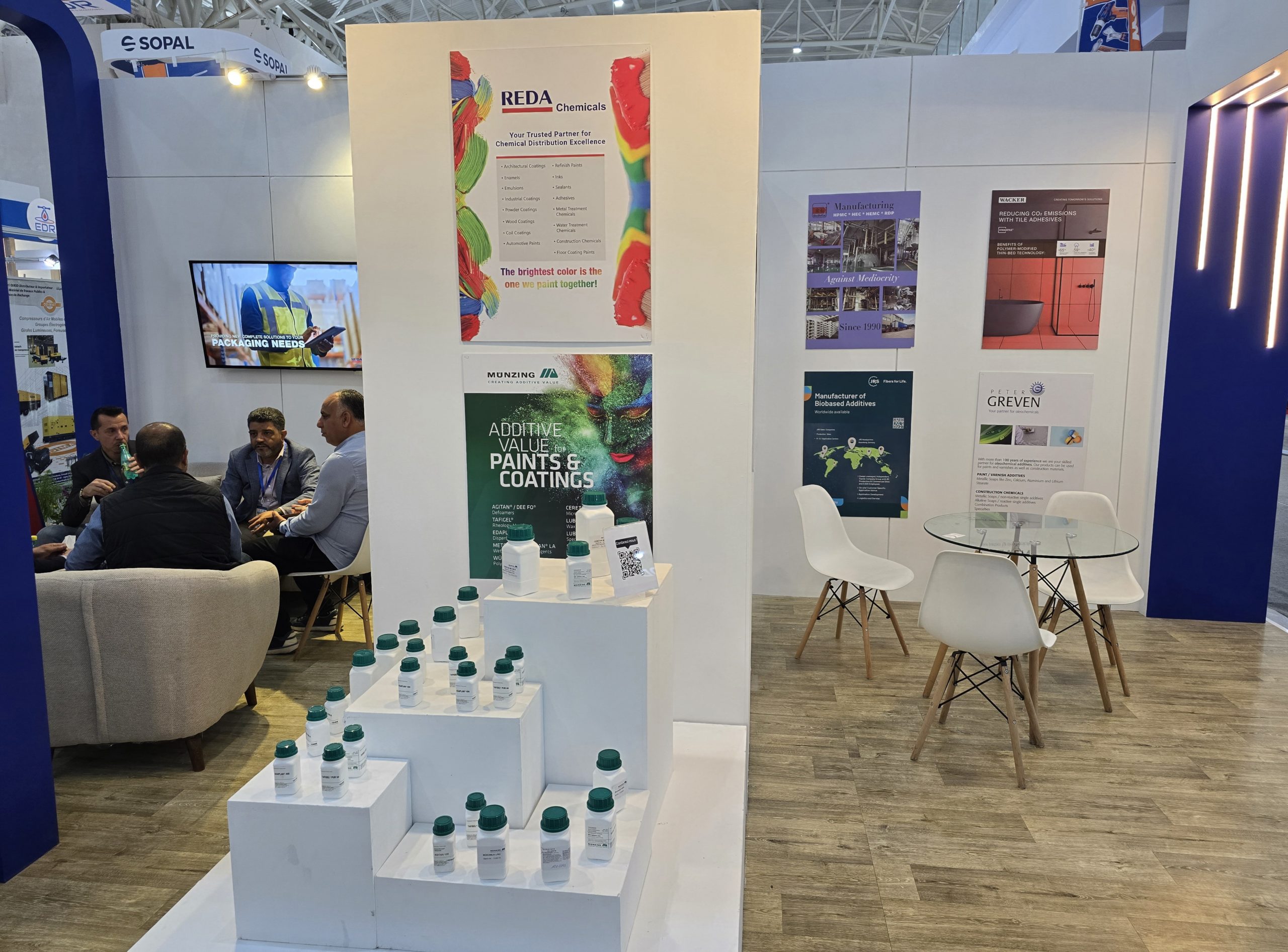 Trade show booth with white stepped platforms displaying many small white bottles with teal caps, colorful posters on the back wall, and people seated nearby.