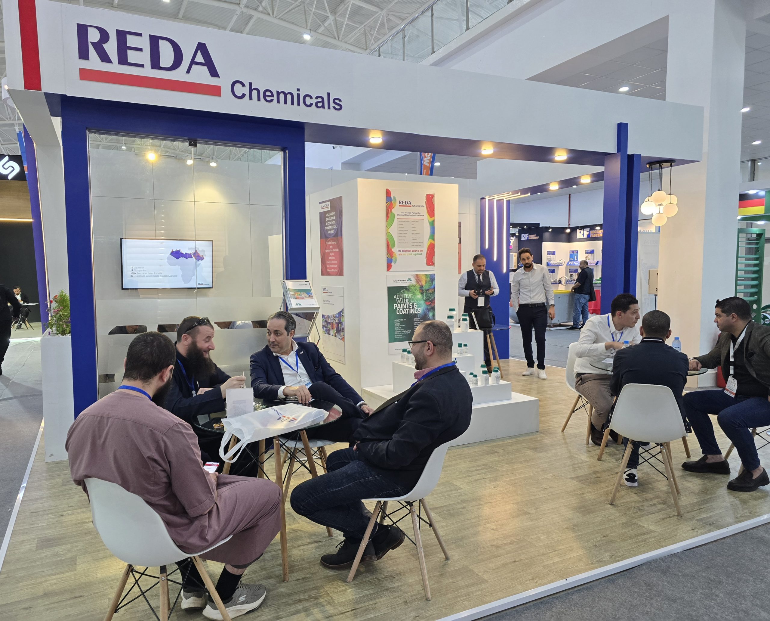 Trade show booth labeled 'REDA Chemicals' with attendees seated at tables in discussion areas inside a bright expo hall