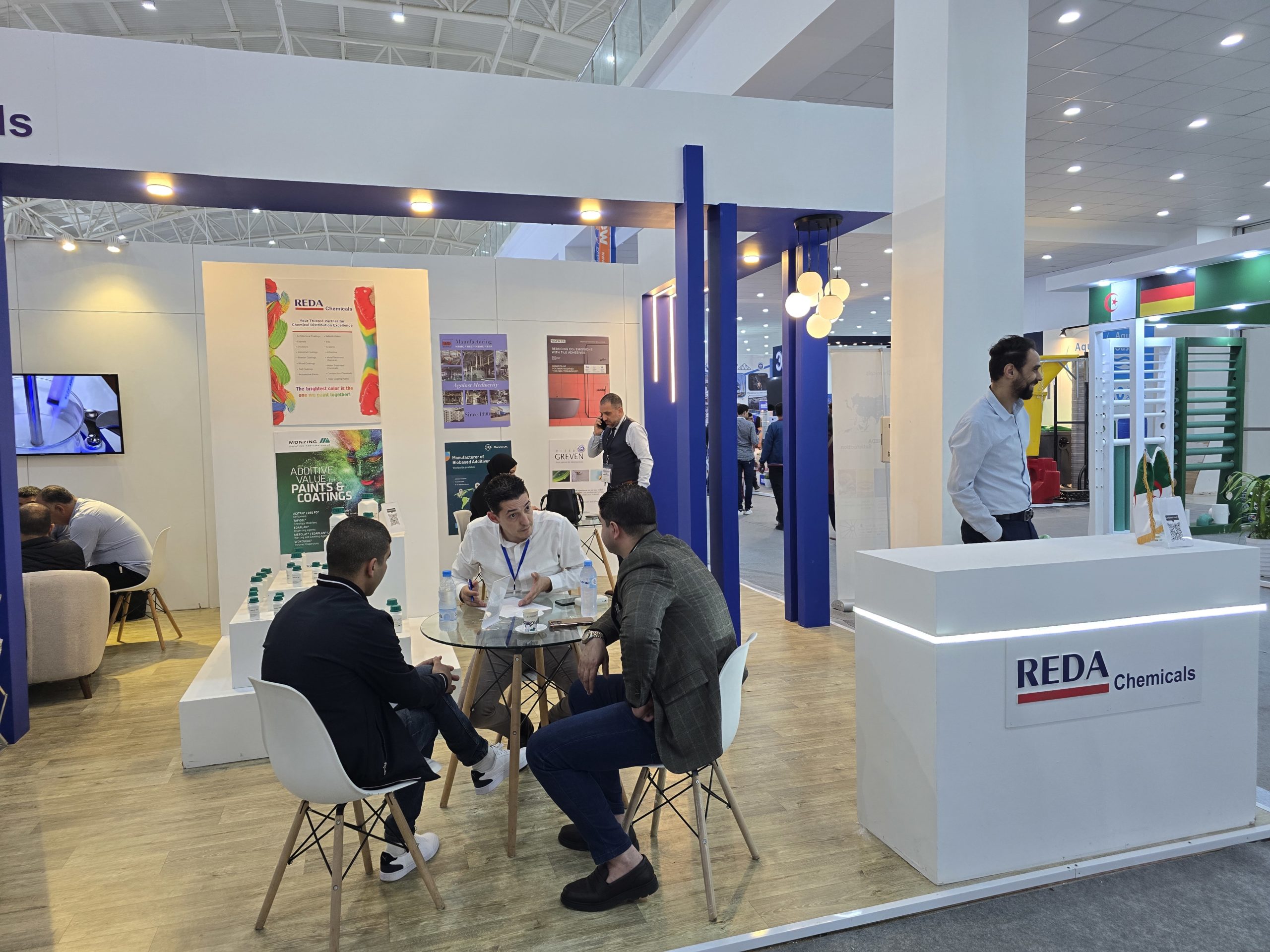 Trade show booth for REDA Chemicals with staff seated at round glass table and posters in background inside a bright exhibition hall.