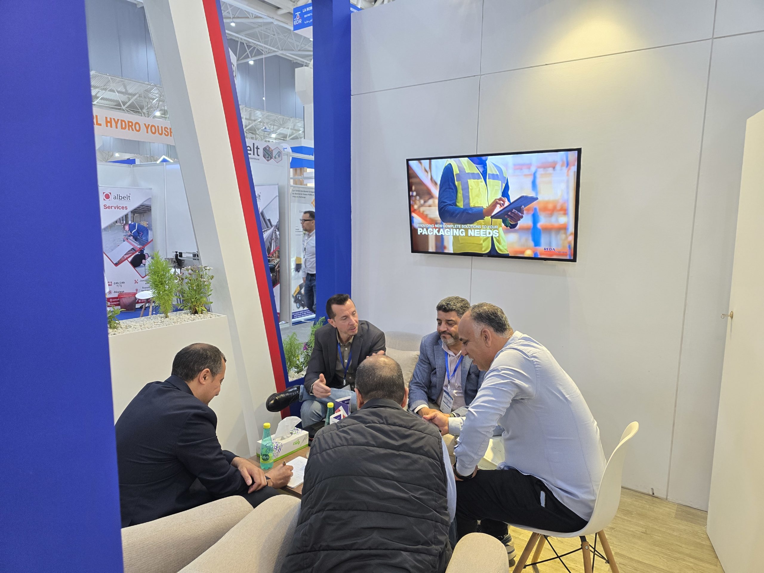 Group of men in business attire seated around a table at a trade show booth, discussing documents, with a wall-mounted screen showing a worker in a yellow safety vest.