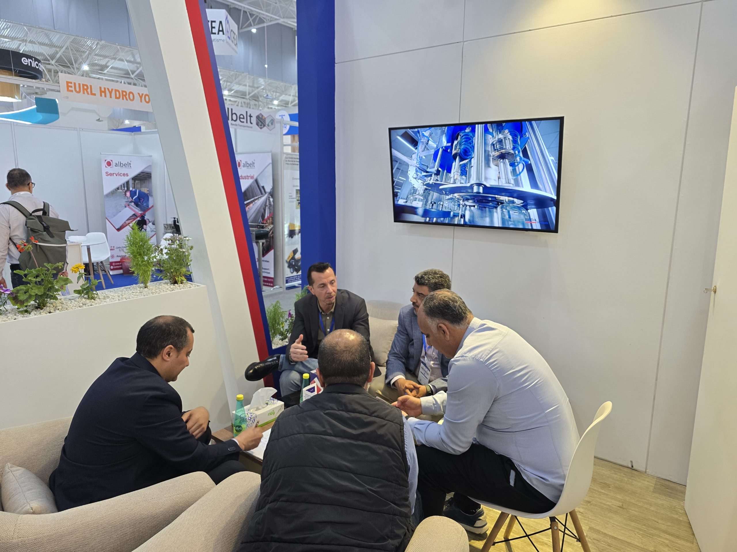 Group of professionals seated around a table at a trade show booth, reviewing documents, with a wall-mounted screen displaying machinery.