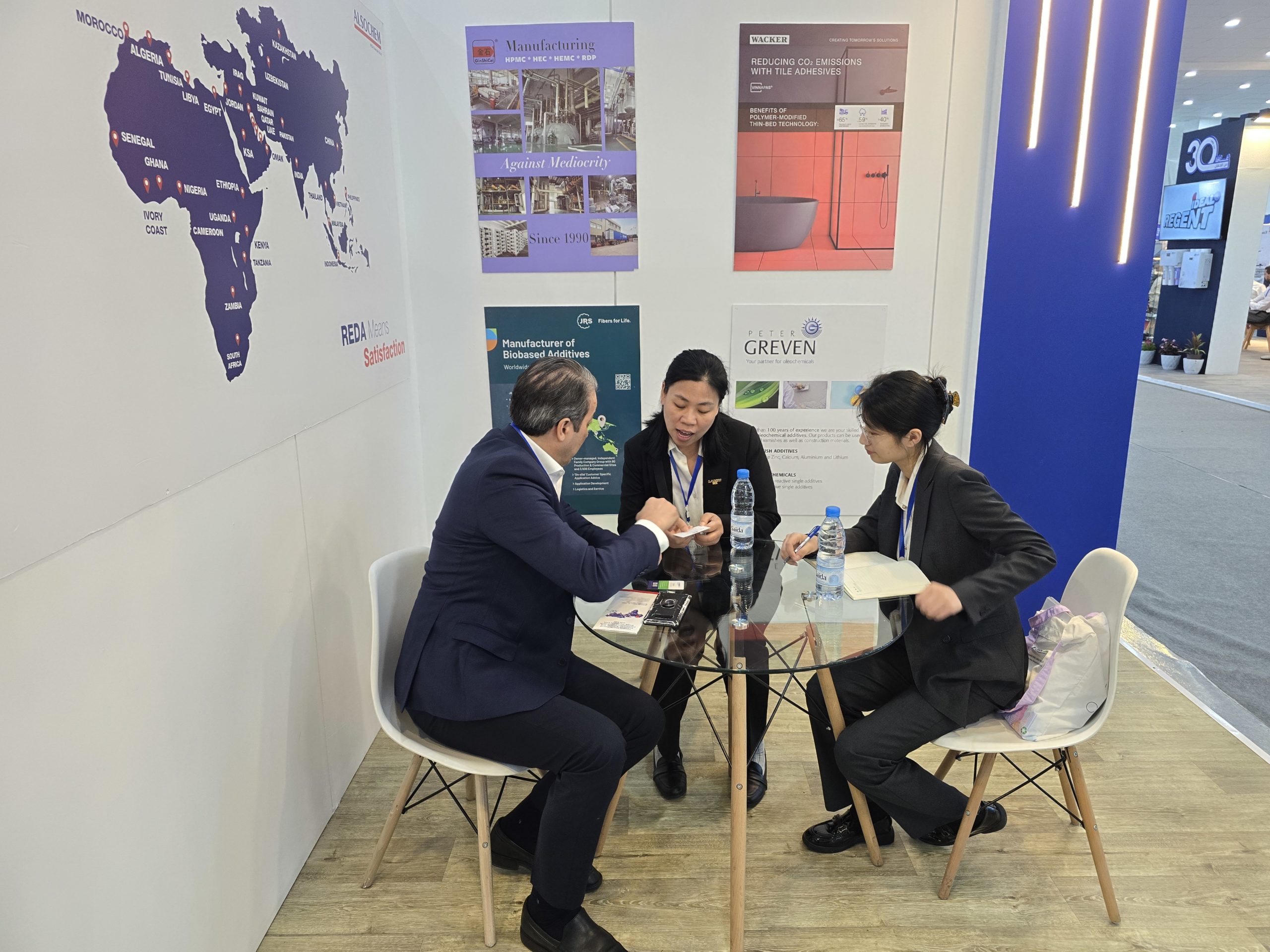 Three business professionals in formal attire sit around a round glass table at an expo booth, discussing documents with posters and a world-map mural on the wall behind them.