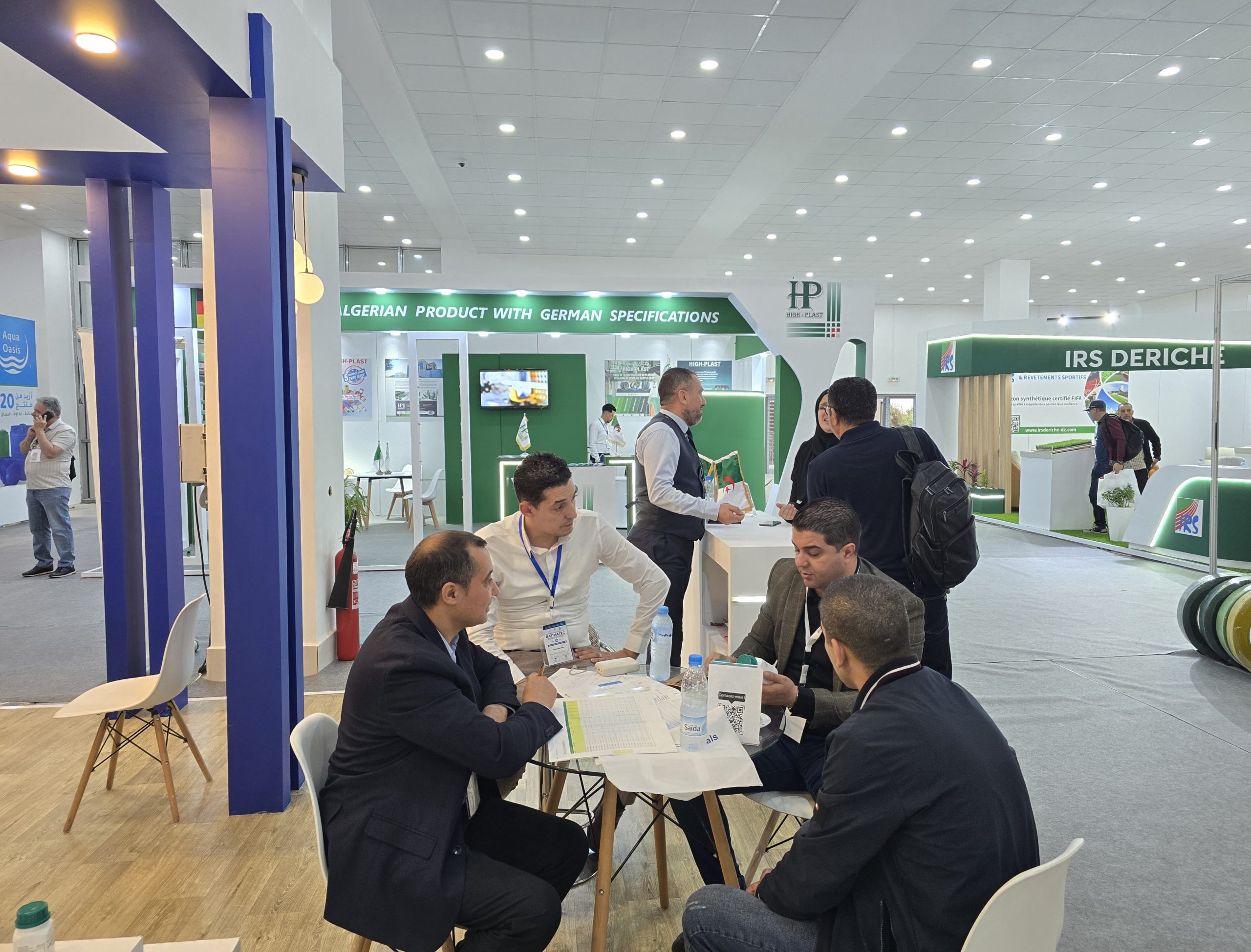 Group of professionals seated at a round table in a trade show, discussing documents with exhibitor booths in the background.