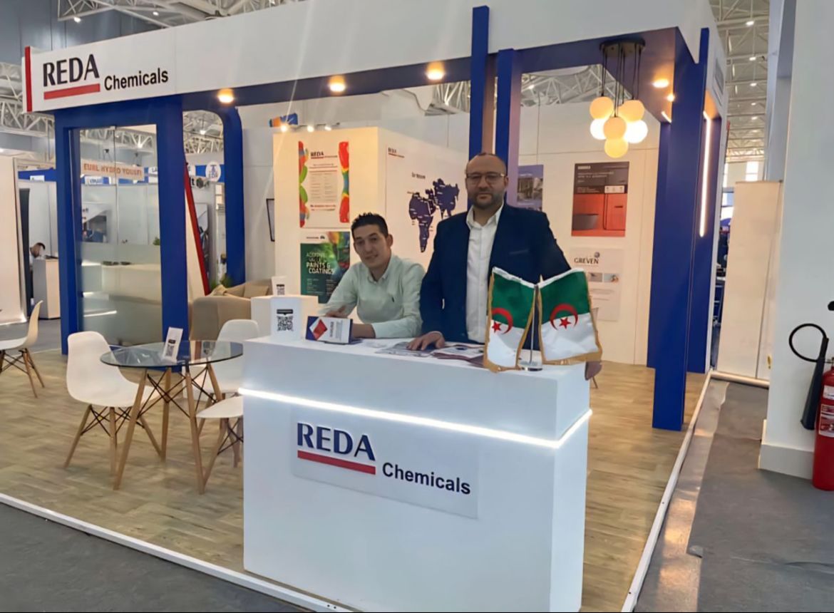 Trade show booth for REDA Chemicals with two men at a white reception desk; Algerian flags on the desk, posters and blue structural frames in the background.