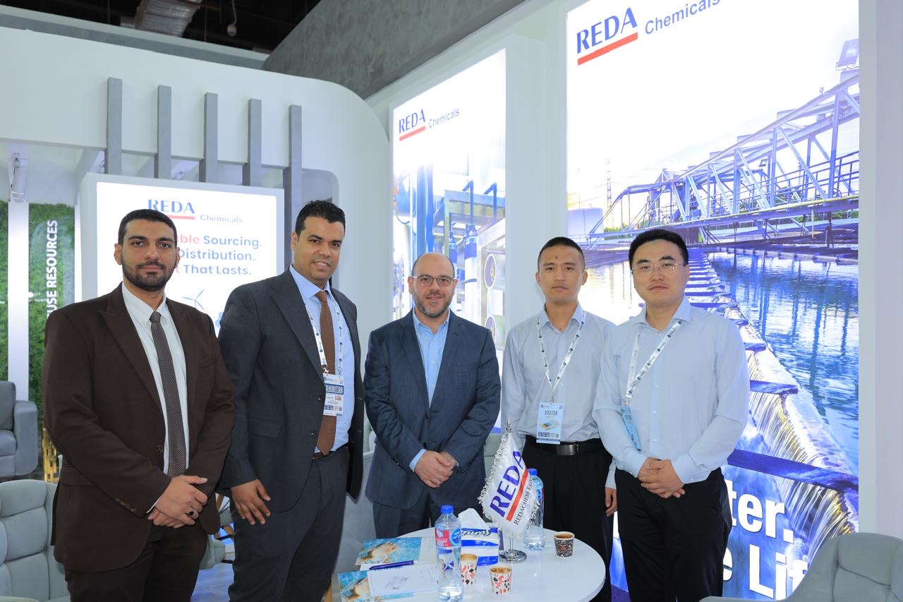 Five men in business attire stand at a REDA Chemicals trade show booth with a blue industrial backdrop behind them.