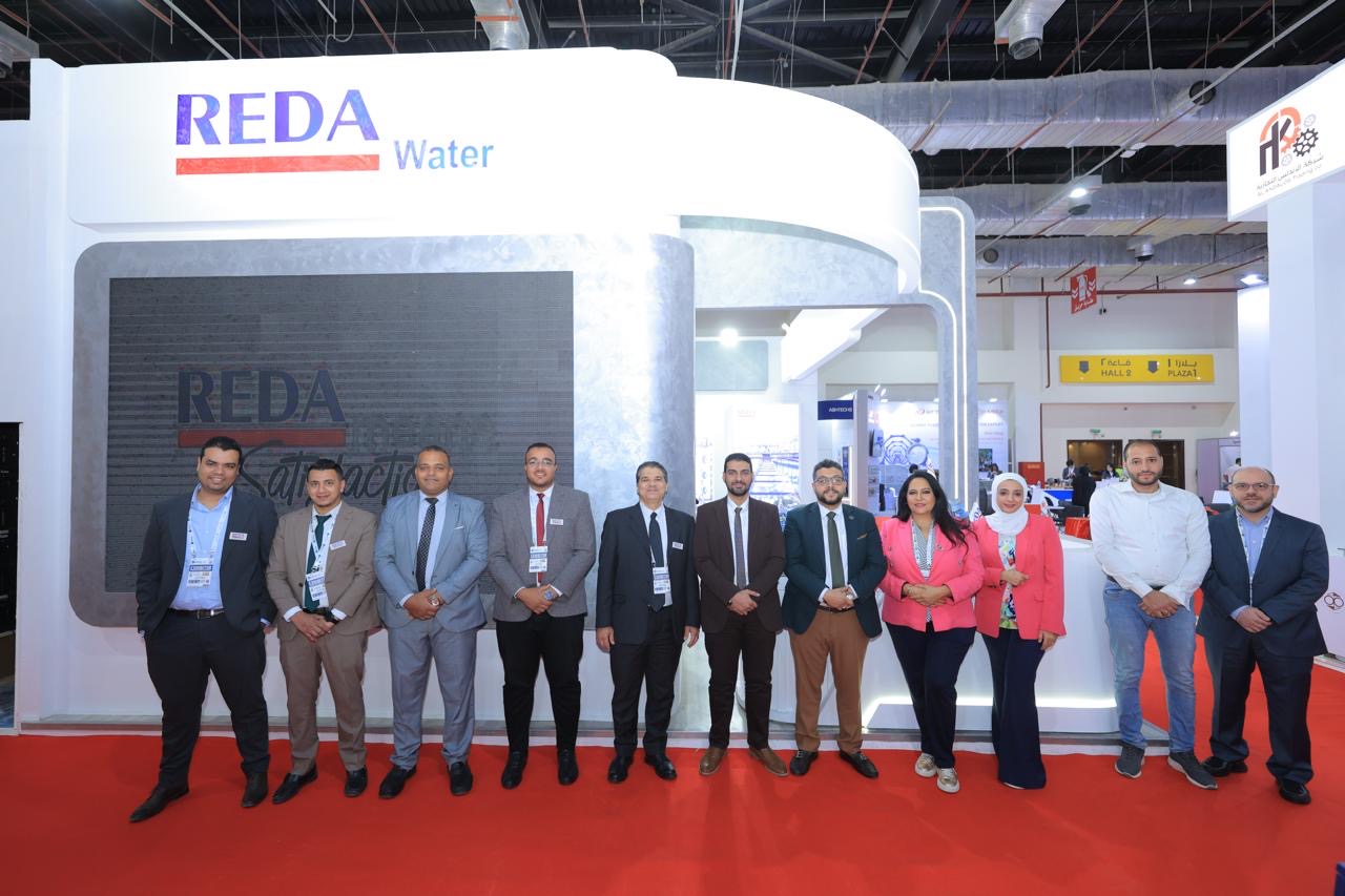 Group of professionals in suits posing for a photo at a REDA Water trade show booth with a red carpet behind them Dated expo backdrop behind the crowd