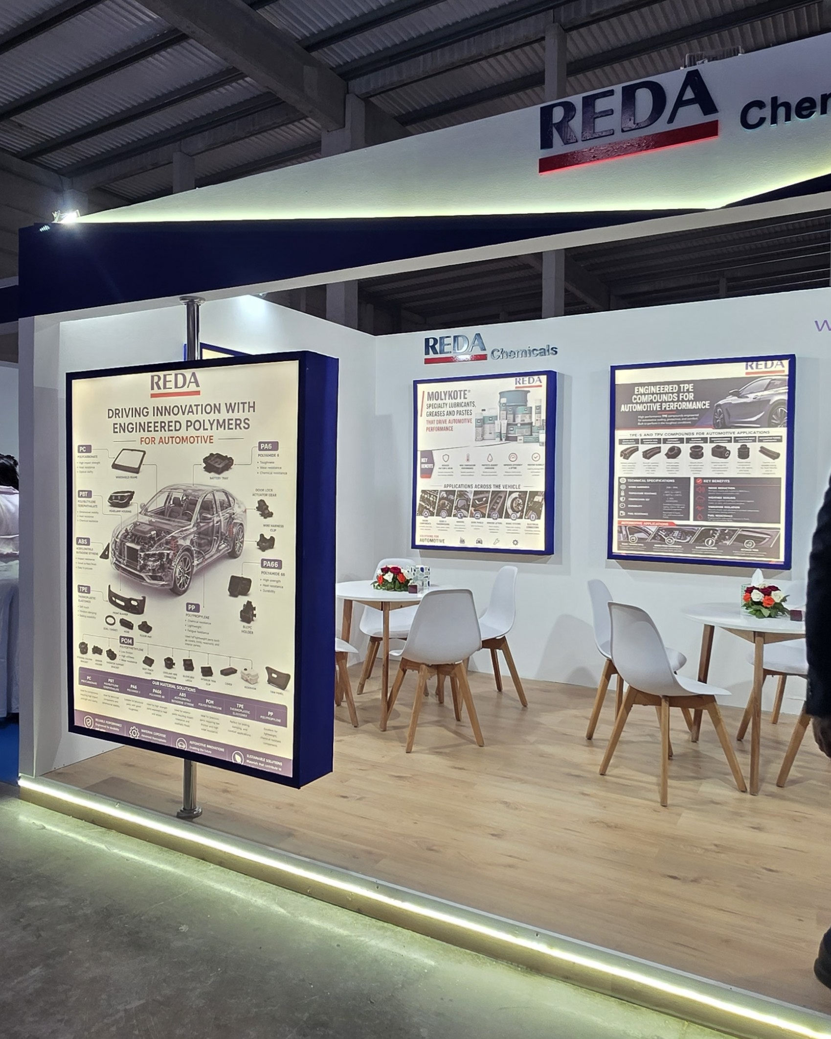 Exhibition booth with white walls and large posters showing REDA Chemicals automotive information, surrounded by white chairs and wooden tables under a metal ceiling pillared display area.