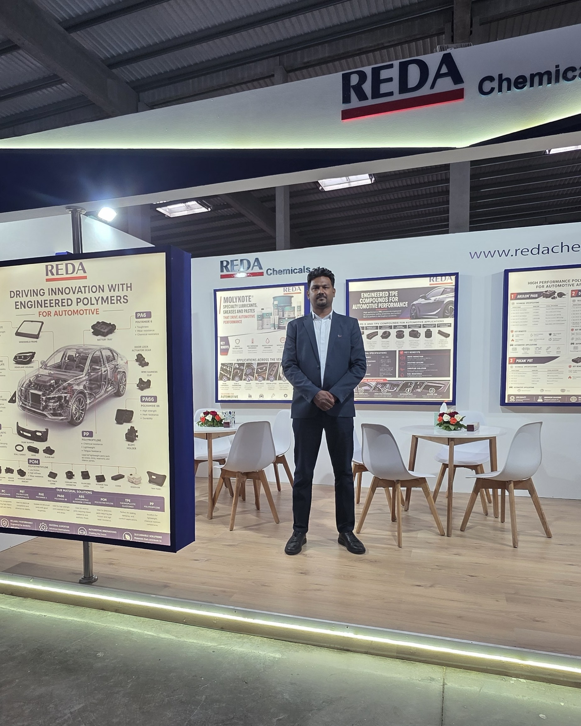 Man in a blue suit standing in front of a REDA Chemicals exhibition booth with large posters and a seating area in the foreground.