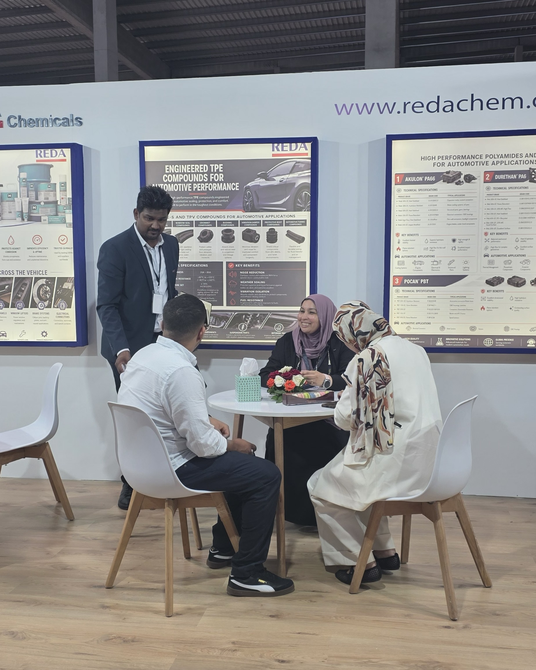 Group of four people seated around a white round table in a trade show booth with large posters on the wall behind them and a tissue box on the table.