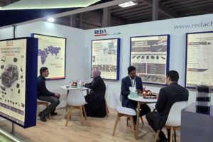 Trade show booth for REDA Chemicals with posters on display and people seated at white tables, discussing products in a modern exhibition hall.