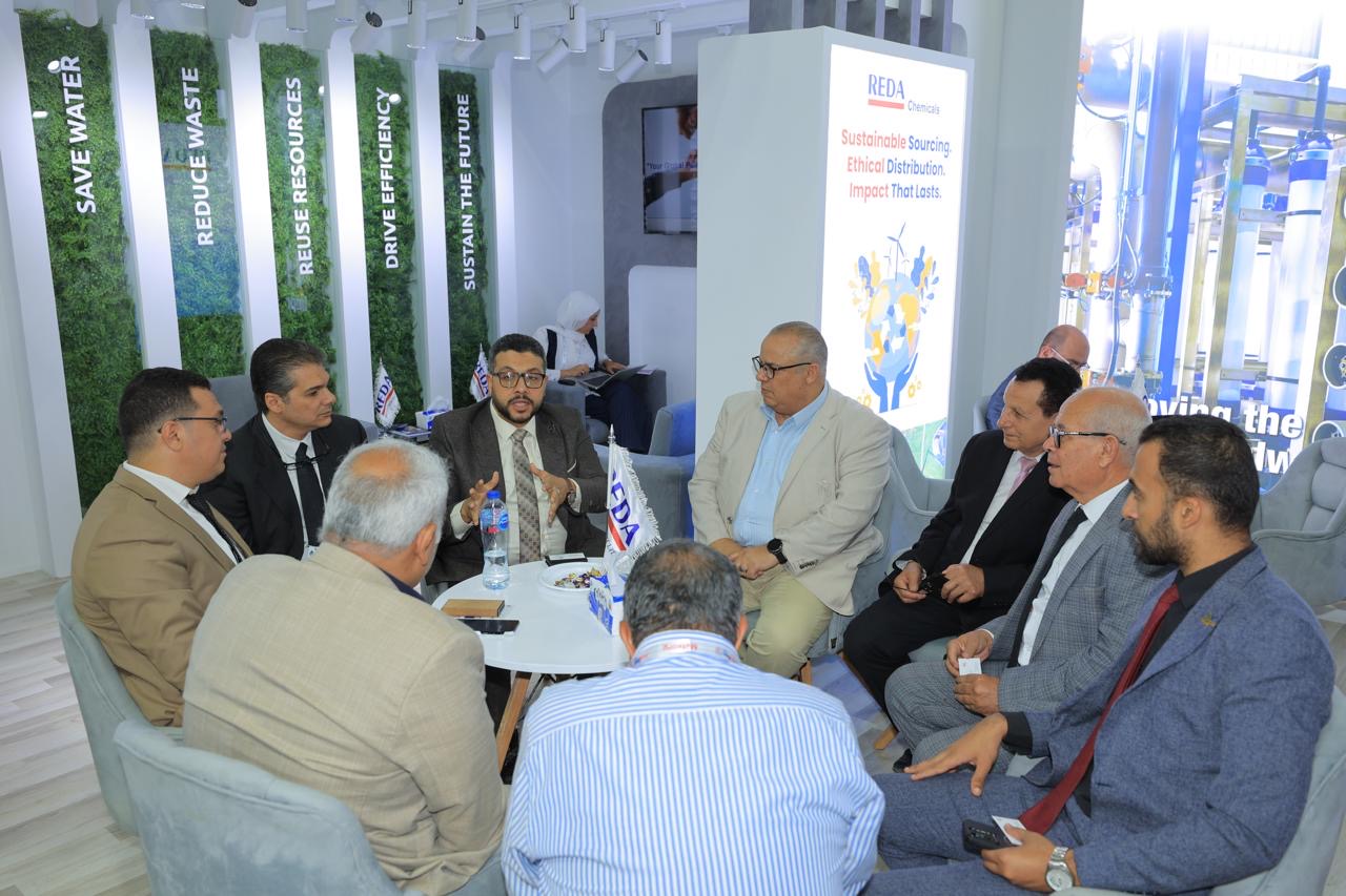 Group of professionals in suits seated around a round white table, having a discussion at a trade-show booth with REDA Chemicals branding and sustainability banners in the background.