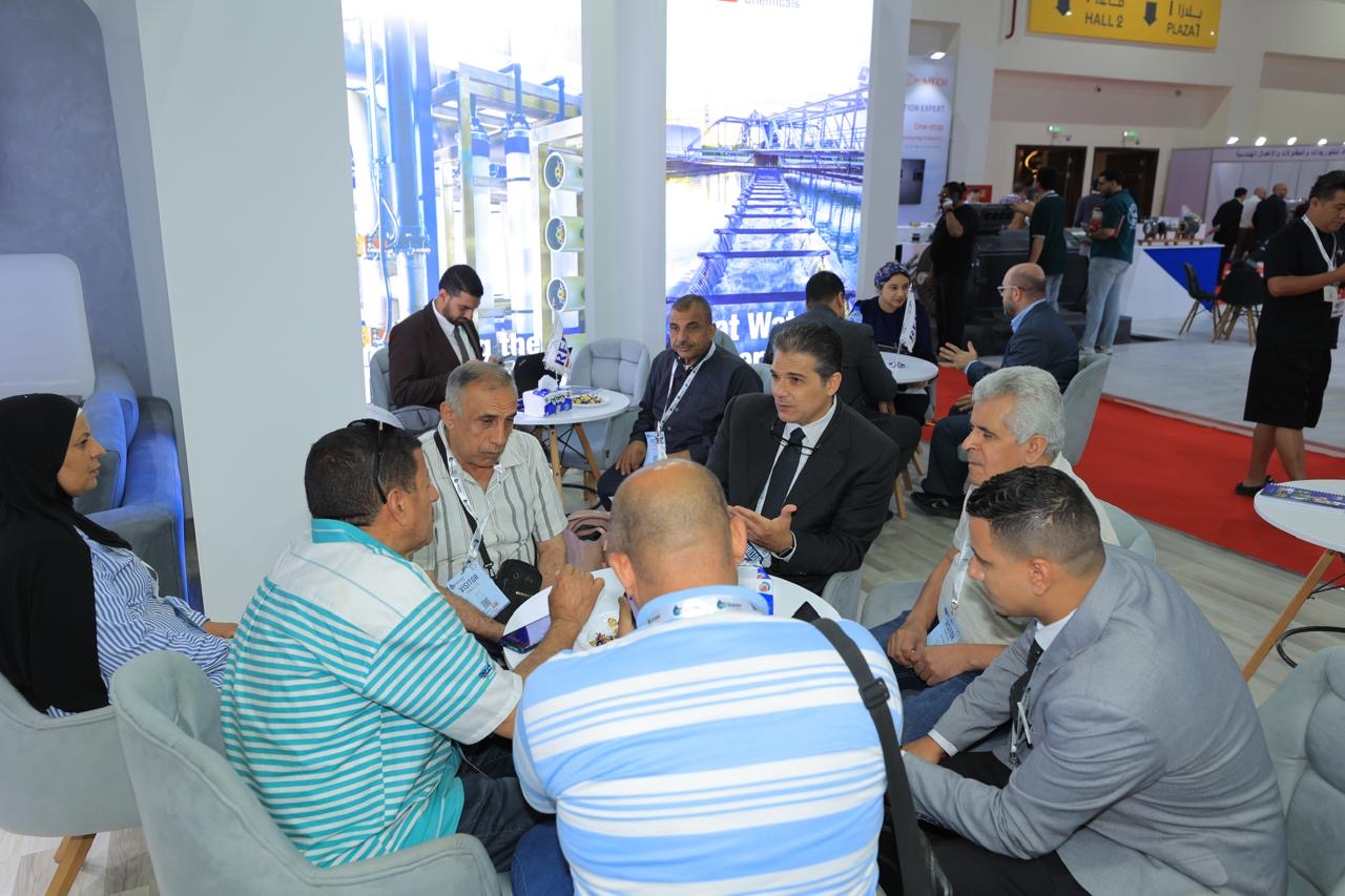 Group of people sit around a small round table at a conference booth, discussing and wearing lanyards in a busy expo hall.
