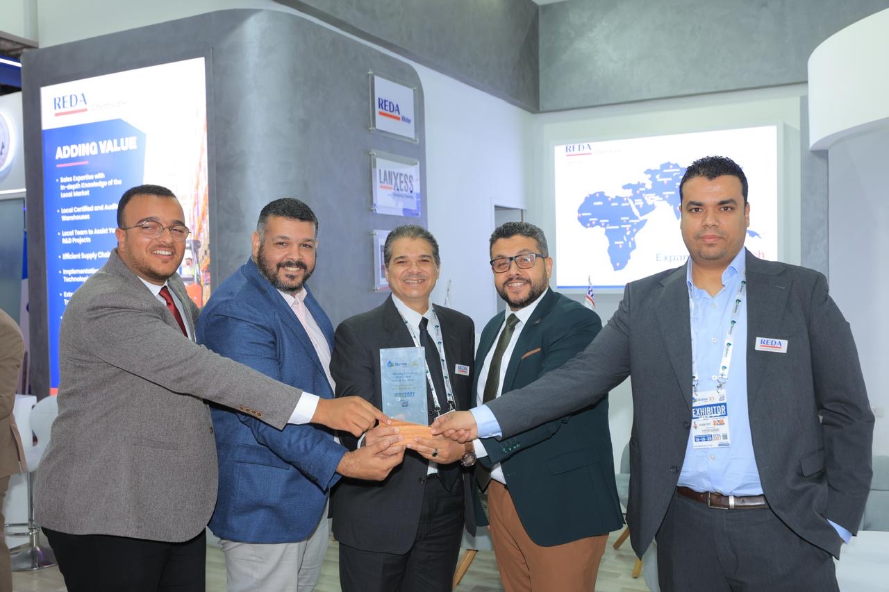 Five men in business attire toast and hold a glass award at a trade show booth.