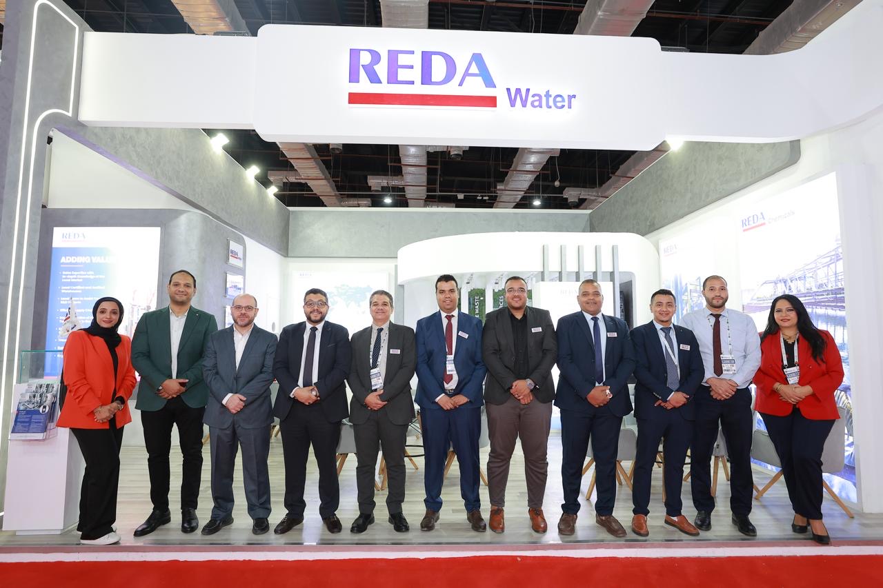 Group of professionals in business attire posing at a REDA Water trade show booth under a bright sign reading REDA Water on top of the booth.