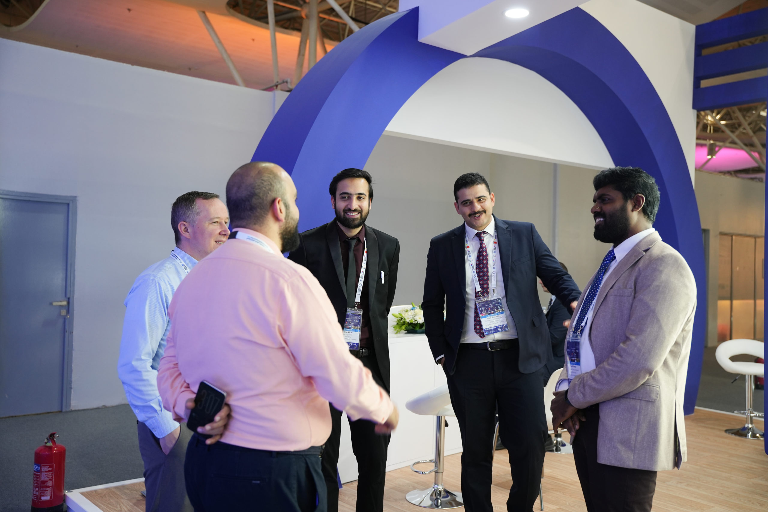 Group of professionals in suits chatting at a conference booth with lanyards and a blue curved exhibit behind them.