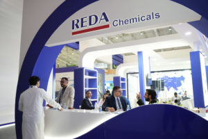 Trade show booth with 'REDA Chemicals' sign, blue and white curved structure, and people in business attire conversing nearby.