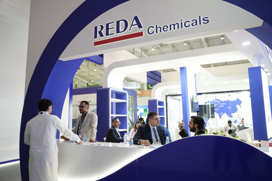 Trade show booth with 'REDA Chemicals' sign, blue and white curved structure, and people in business attire conversing nearby.