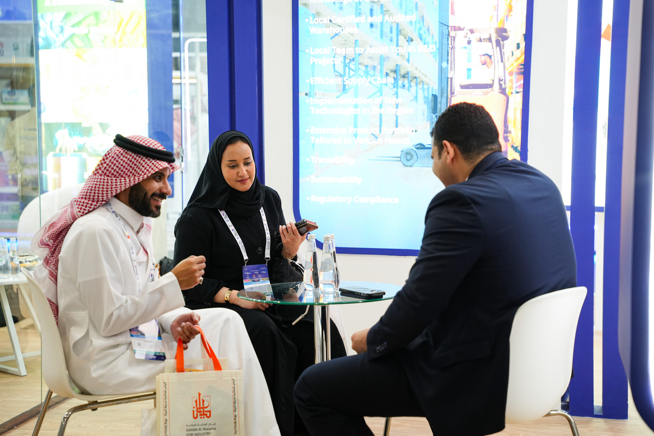 Three professionals sit around a glass table at a trade-show booth, conversing with a man in a dark suit.