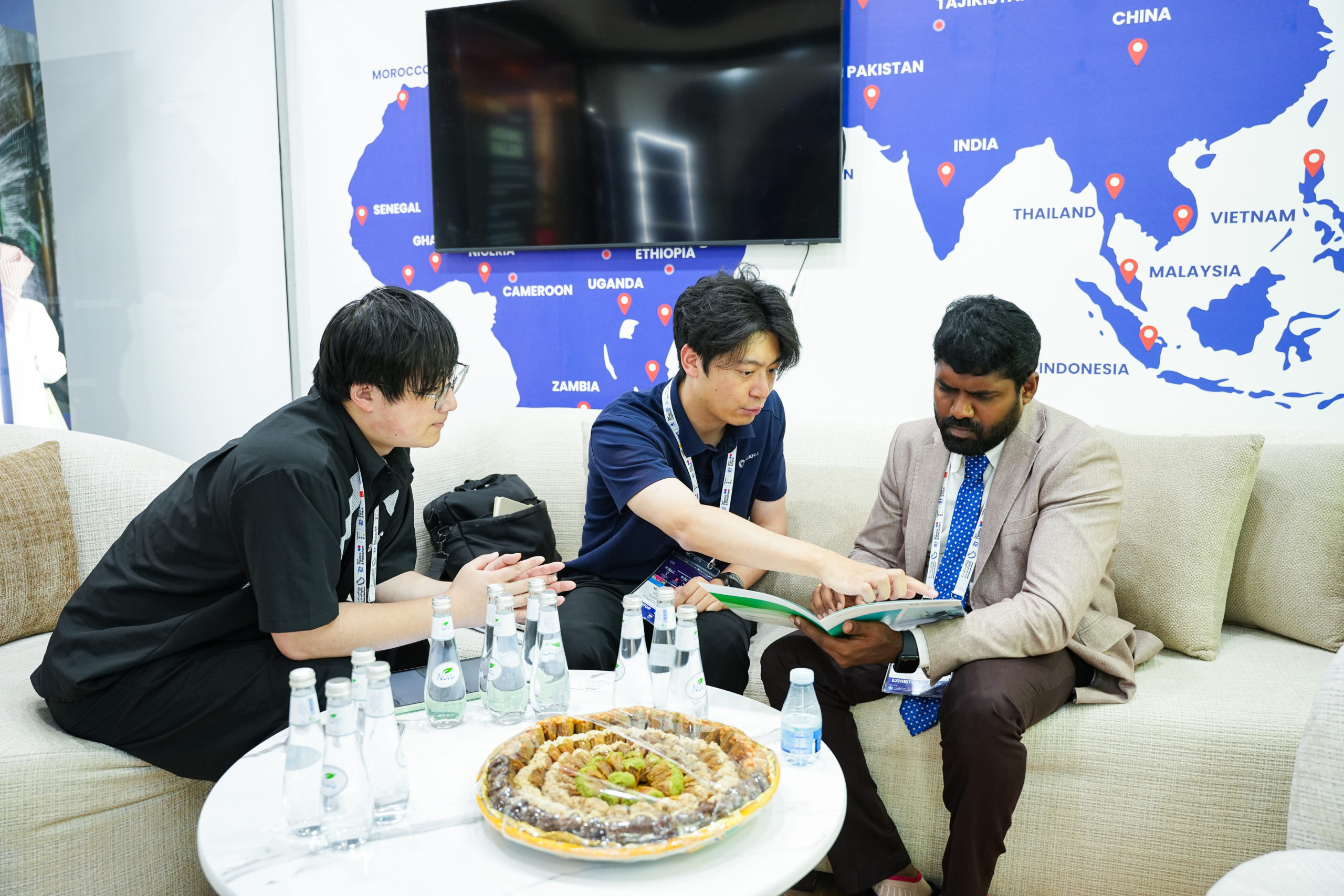 Three men in business casual clothes sit on a beige sofa around a table with bottled water and pastries, discussing a document, with a blue world map backdrop behind them.