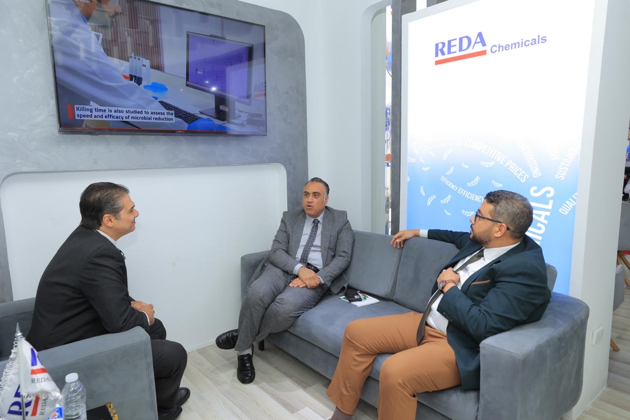 Three men in business attire sit on a gray couch in a modern REDA Chemicals booth, discussing with a lab scene displayed on a wall screen above them.