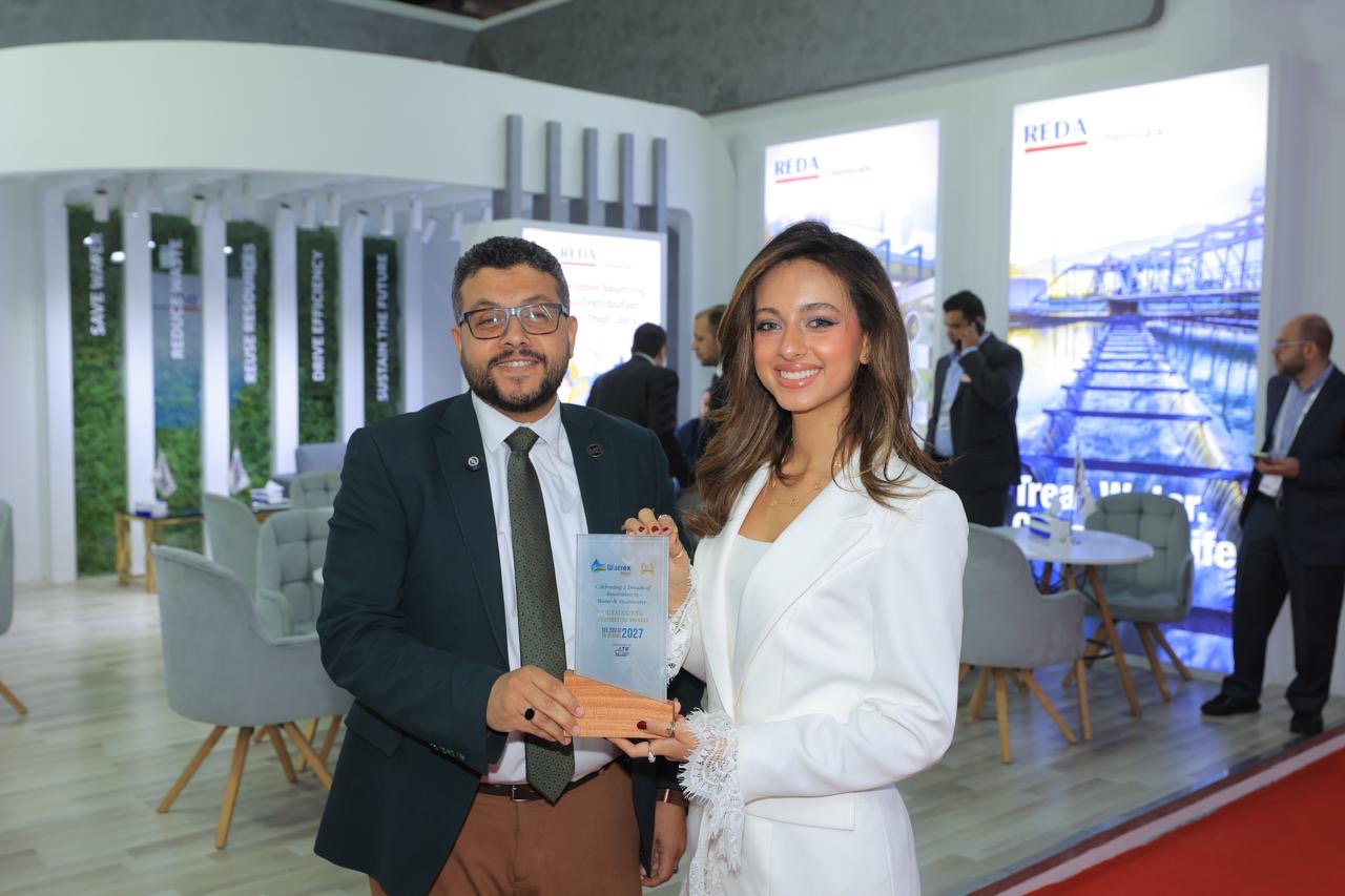 Two professionals in business attire exchange a glass award at a conference booth, smiling at the camera.