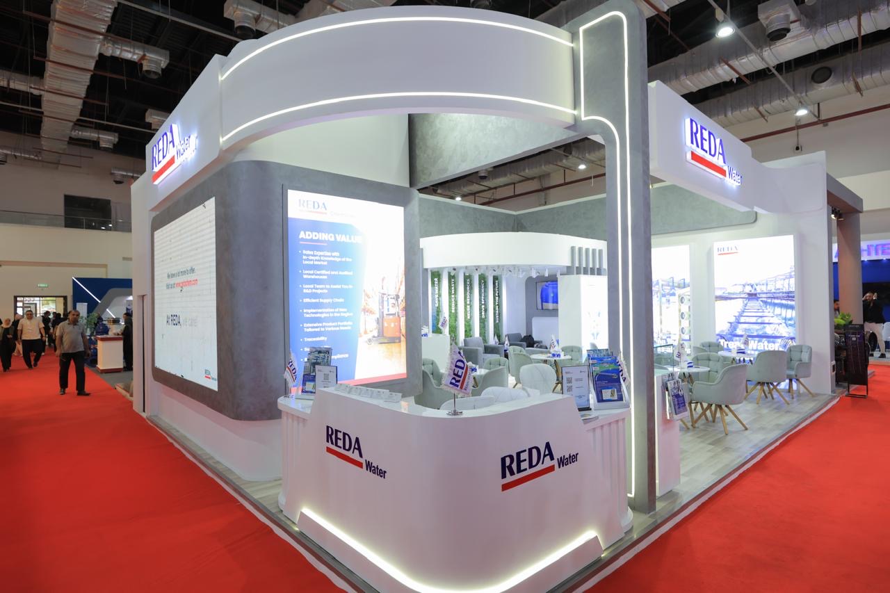 Trade show booth for REDA Water with a white curved arch, reception desk, seating area, and large information screens.”,