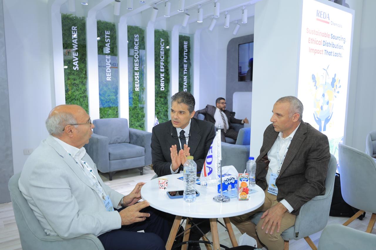 Three men in suits sit around a small round table at a sustainable‑living booth, engaged in a discussion.
